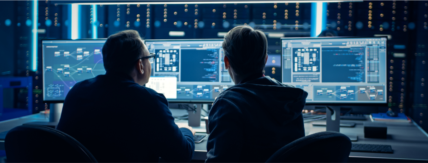 Two male engineers are analyzing code on multiple screens in a computer lab