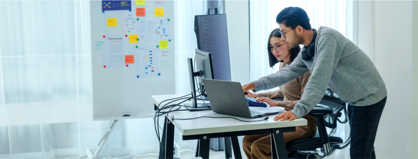 A male and a female engineer are working on at a desk with three screens in an office setting