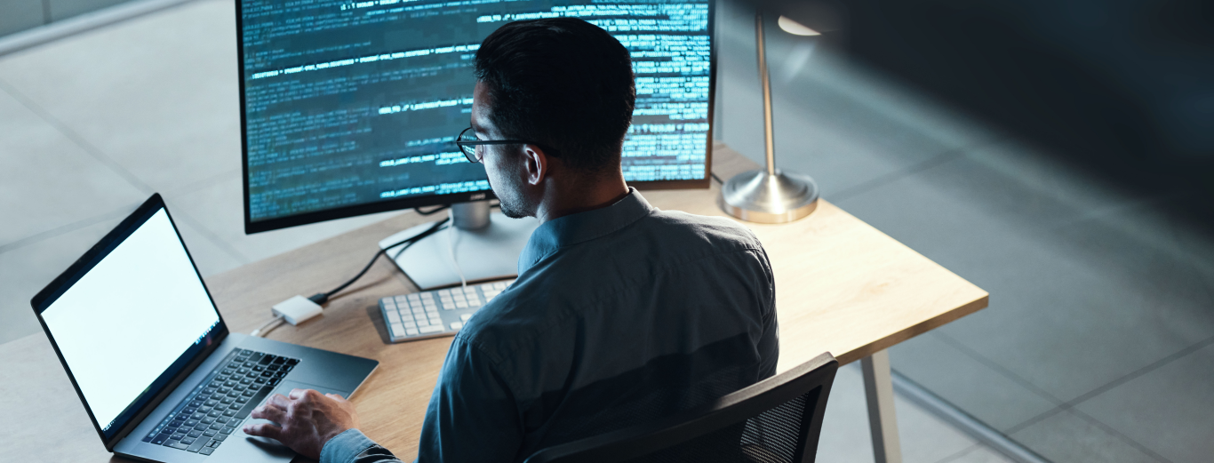 A male IT professional is working on computer screens to develop software code