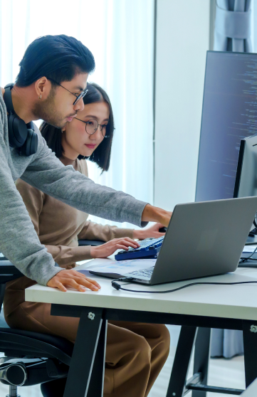 A male and a female engineer are working on at a desk with three screens in an office setting