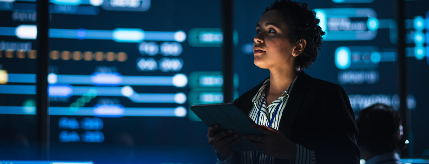 A female IT professional is analyzing data on large screens in a server room