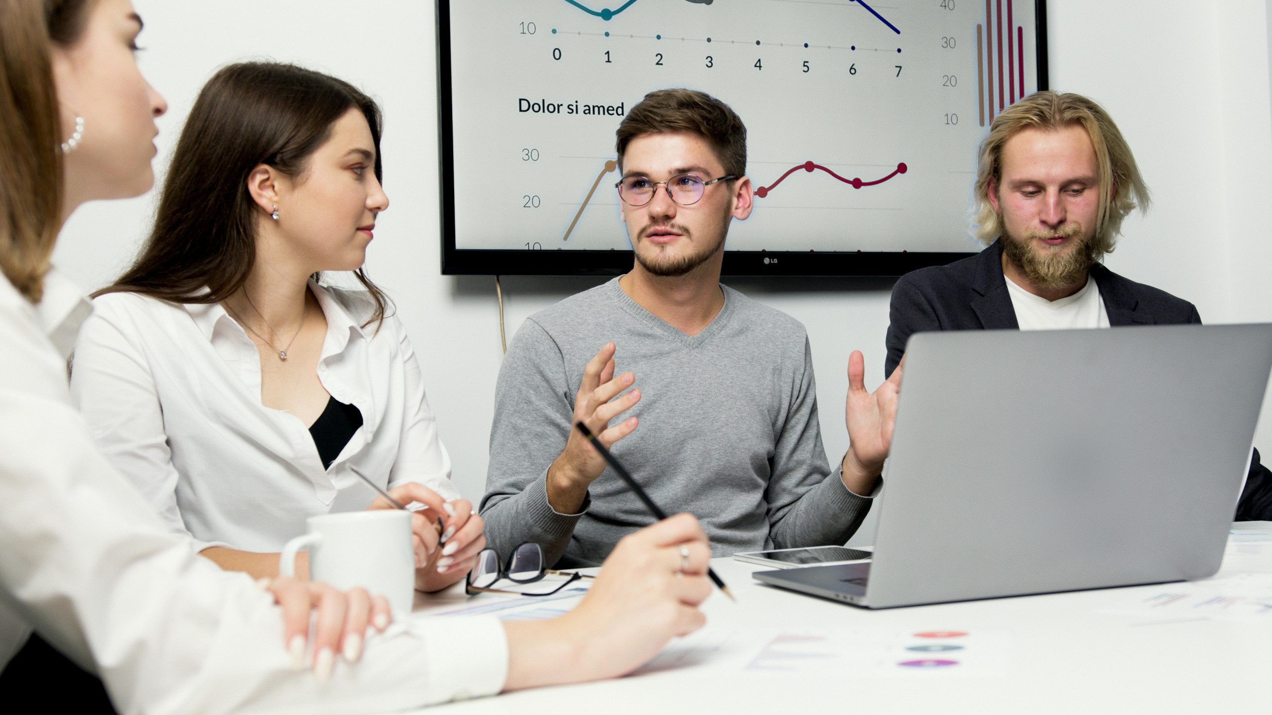 A group of four professionals in a modern office meeting, with one man actively presenting to the others while gesturing.